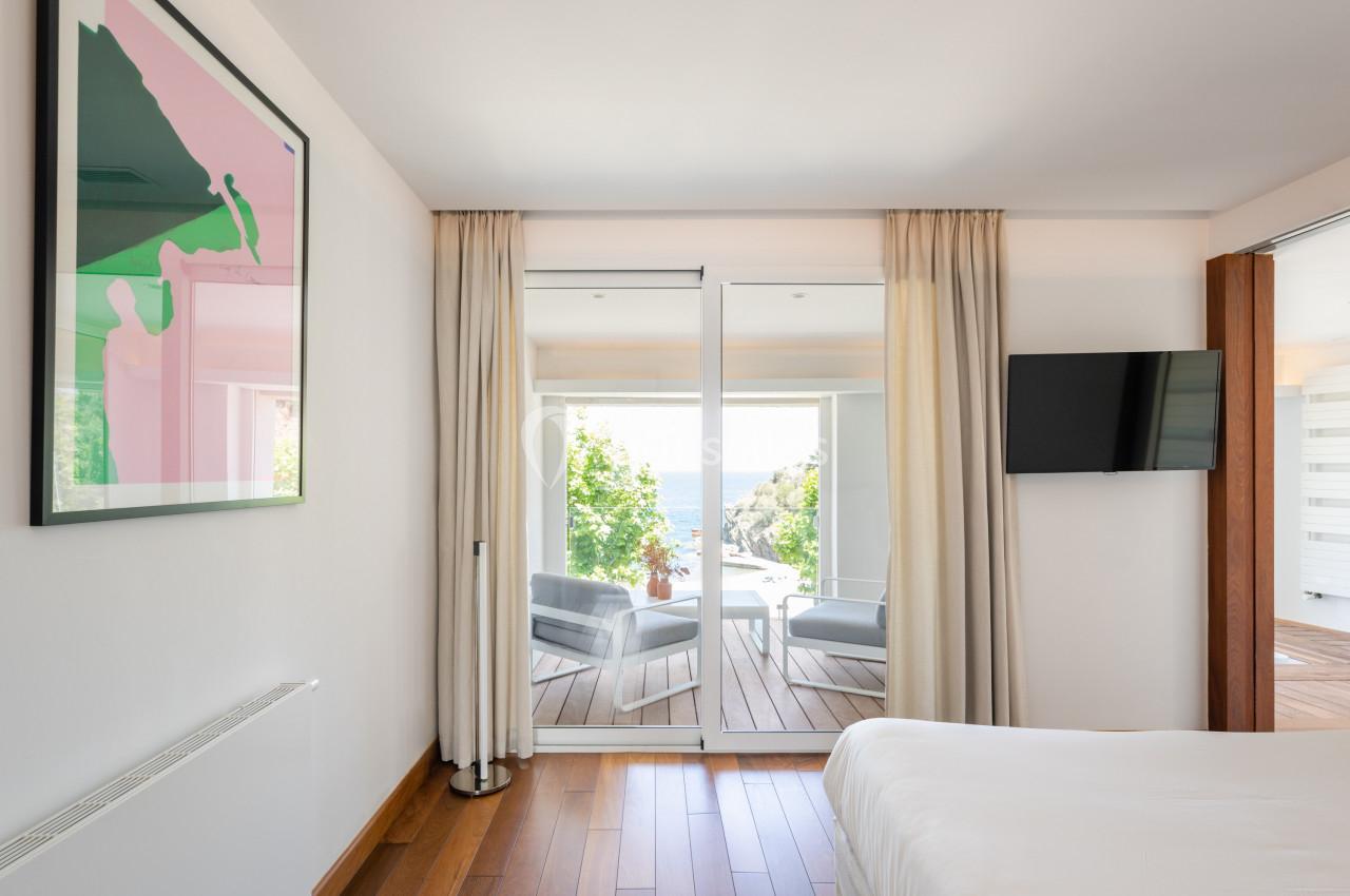 Chambre lumineuse avec lit, tableau mural, télévision et baie vitrée donnant sur une terrasse avec vue sur la mer.