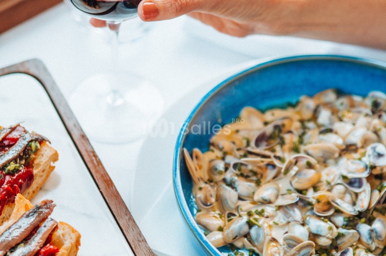 Assiette de palourdes en sauce à côté de tartines garnies, une main tenant un verre de vin rouge.