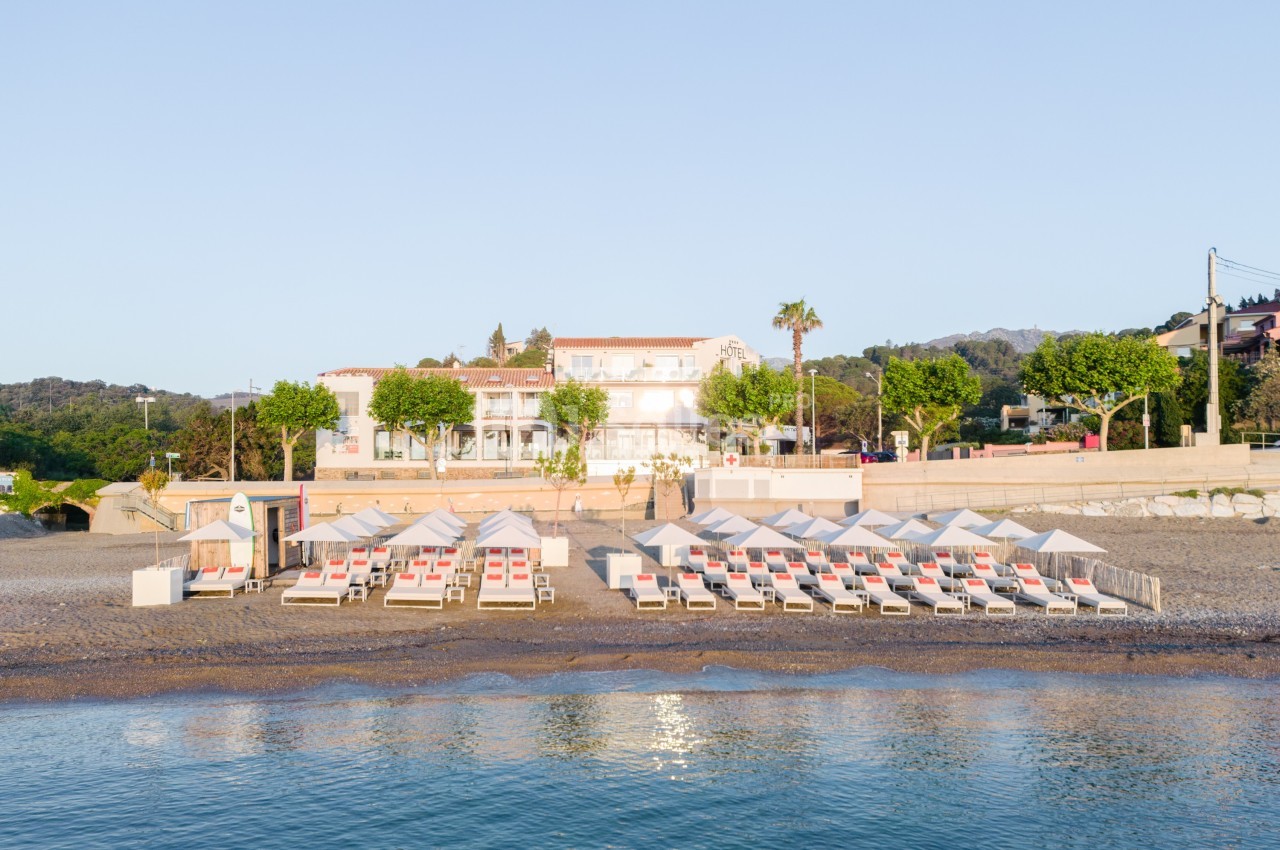Alignement de transats et parasols blancs sur une plage de galets, avec un bâtiment en arrière-plan sous un ciel dégagé.
