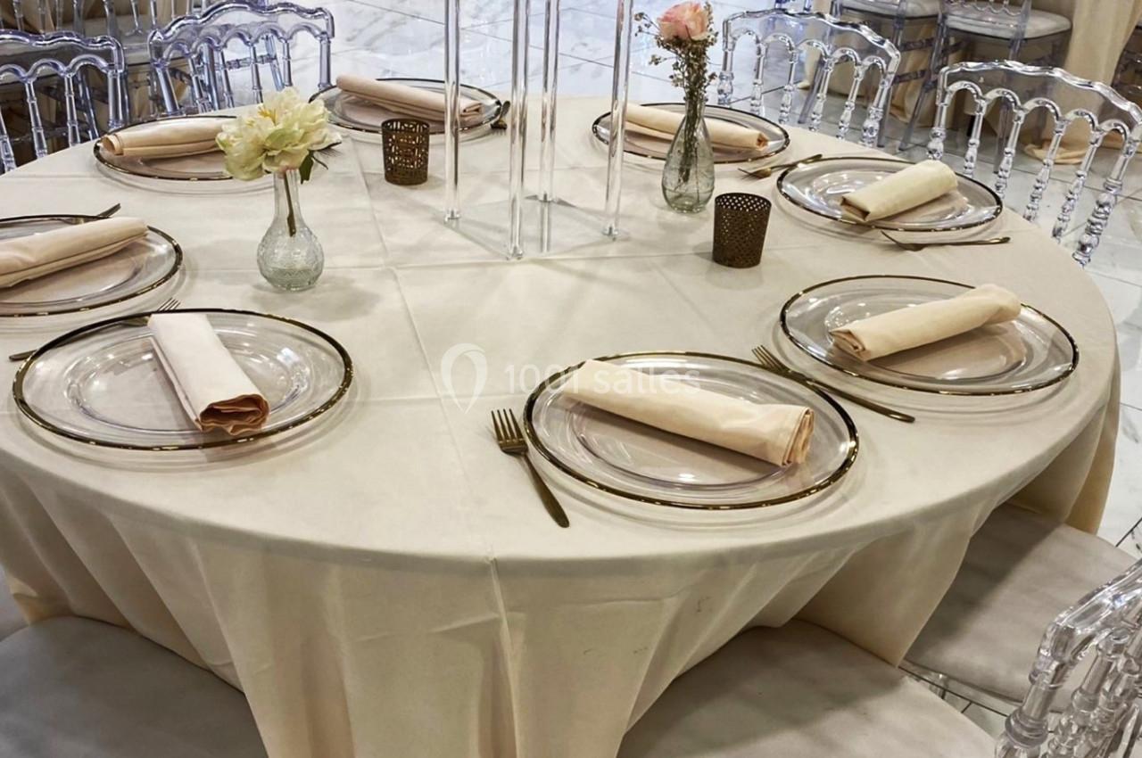 Table ronde élégamment dressée avec nappes blanches, assiettes transparentes, serviettes pliées et décorations florales.
