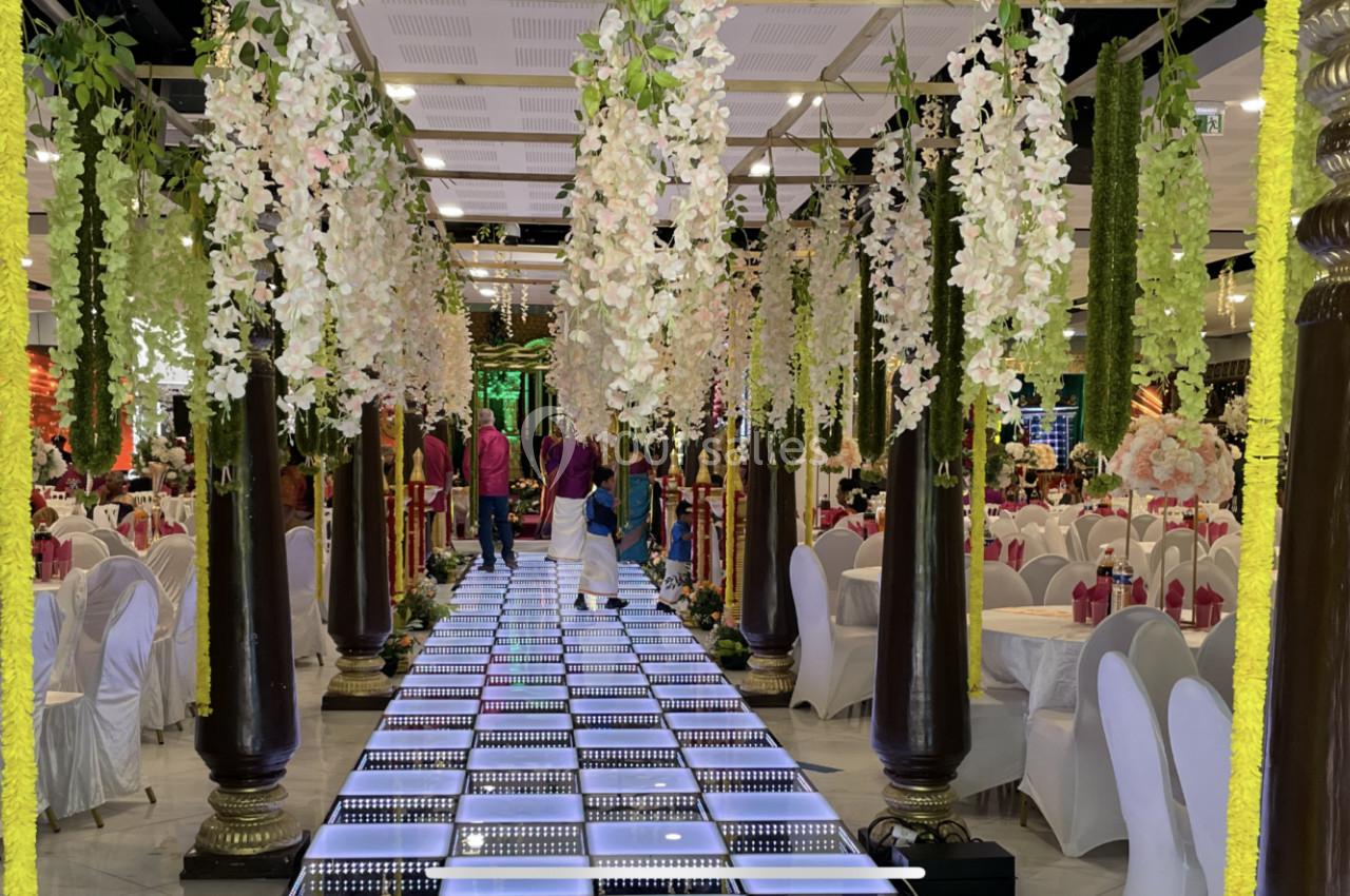 Allée décorée de colonnes, fleurs suspendues et sol lumineux dans une salle de réception ornée de tables dressées.