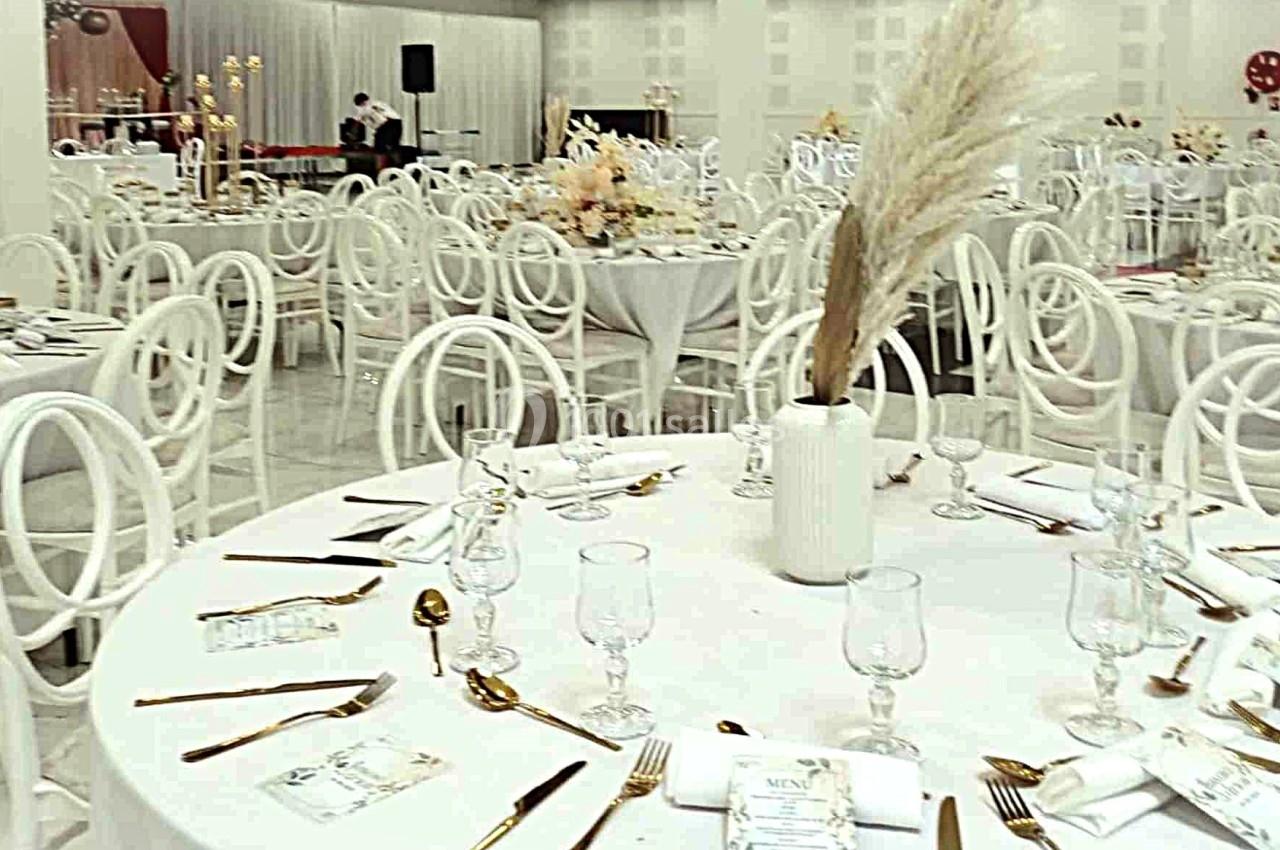 Salle de réception décorée avec des tables rondes dressées, chaises blanches et centre de table floral.