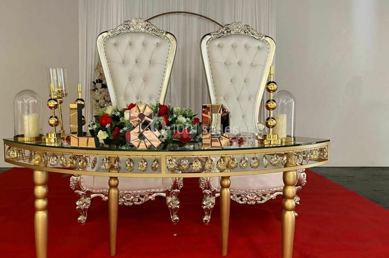 Table décorative dorée avec ornements floraux et bougies, devant deux fauteuils blancs capitonnés sur tapis rouge.