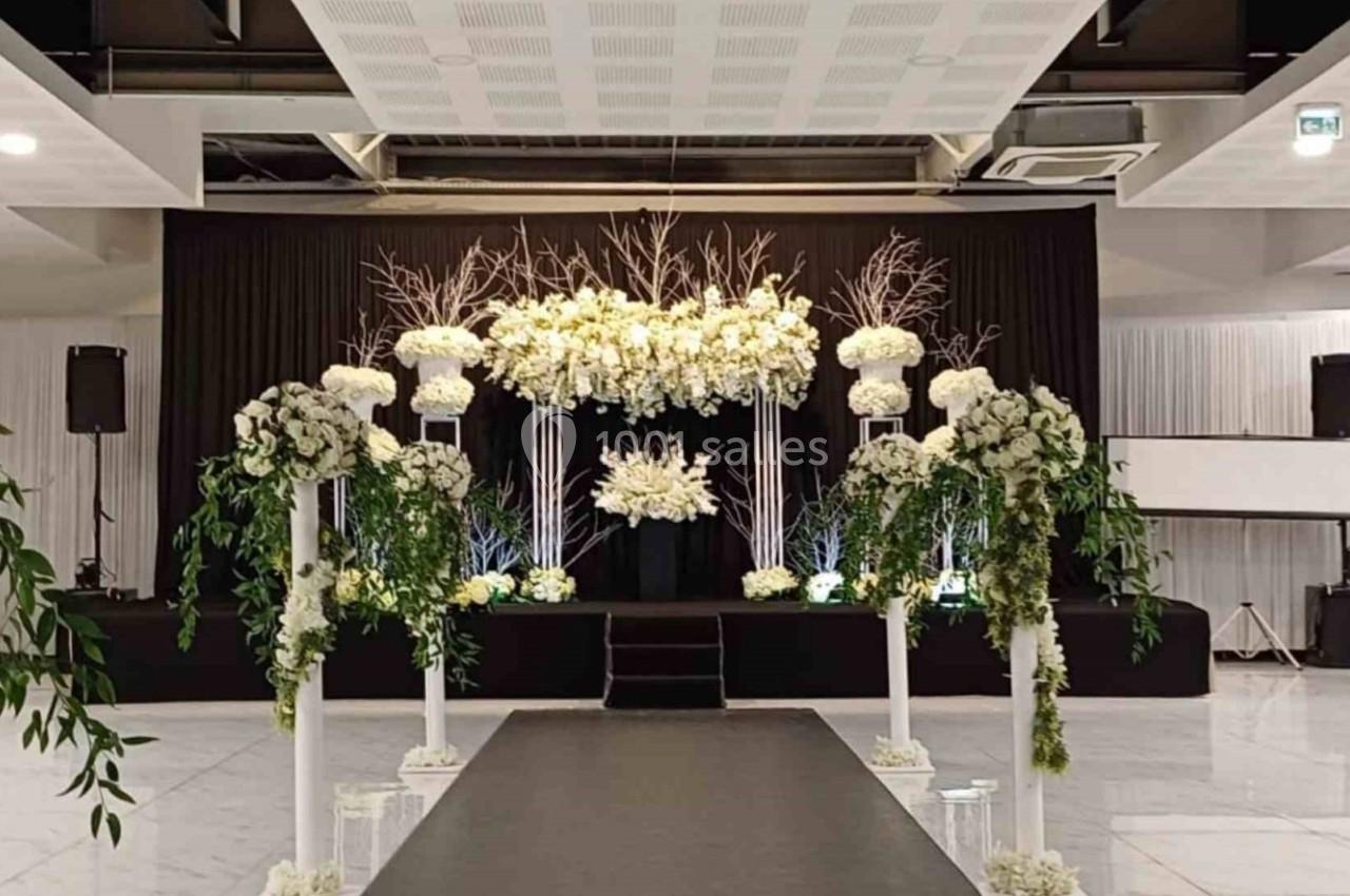 Allée décorée de fleurs blanches et verdure menant à une scène ornée d'arrangements floraux dans une salle de réception.