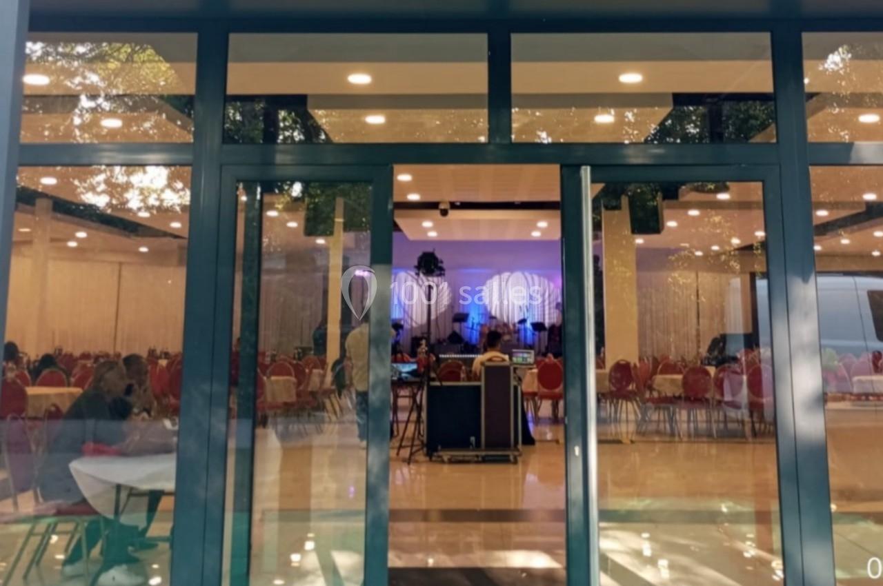 Vue extérieure d'une salle de réception avec des tables dressées et des chaises rouges visibles à travers les baies vitrées.