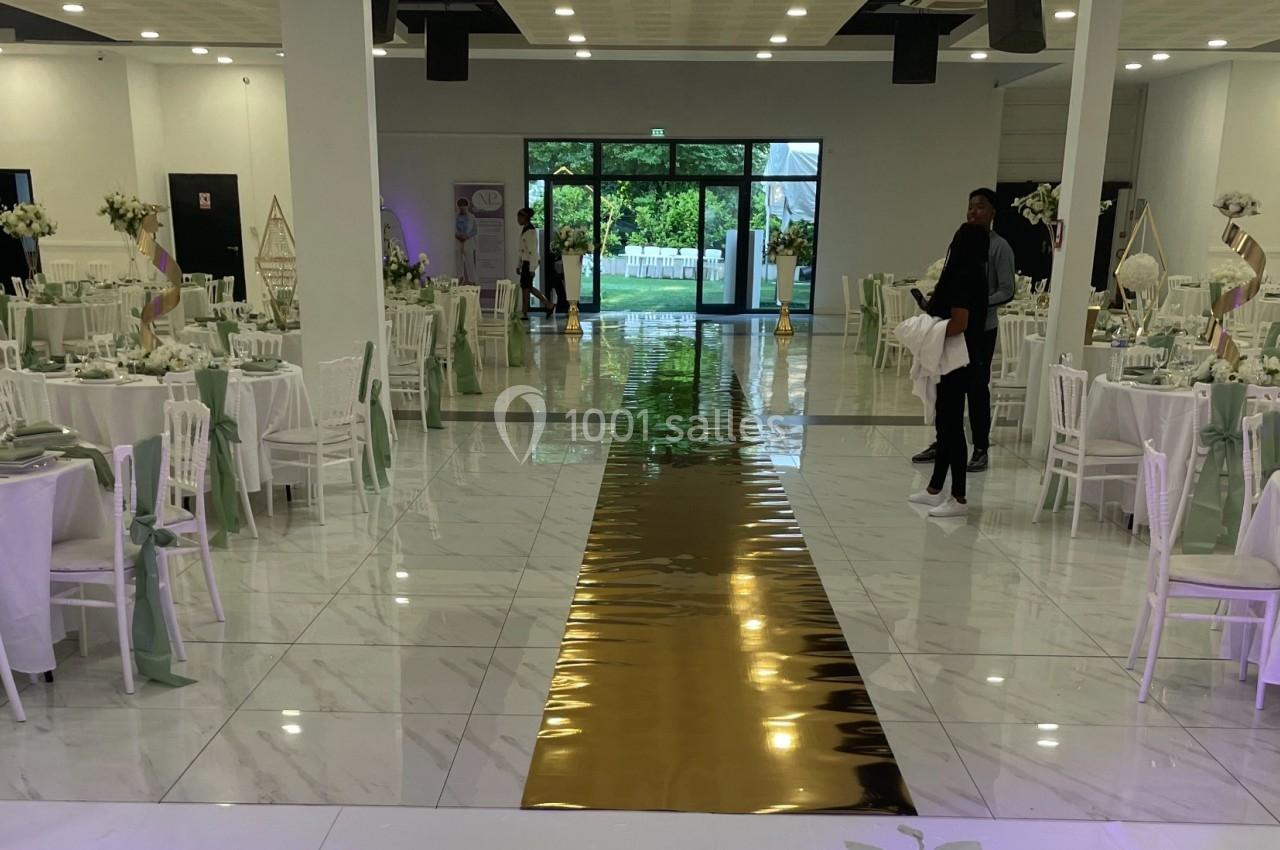 Salle de réception décorée avec des tables rondes, nappes blanches, chaises assorties et une allée centrale dorée.