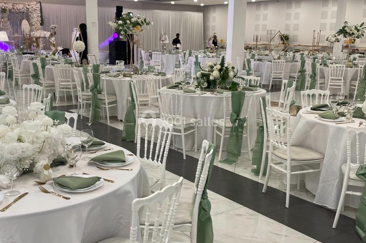 Salle de réception décorée avec des tables rondes, nappes blanches, chaises blanches et rubans verts, ambiance élégante.