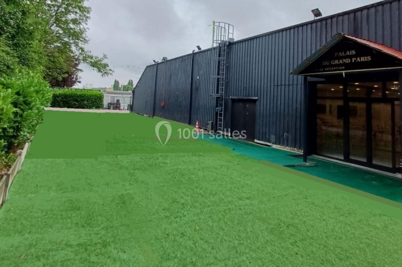 Extérieur d'un bâtiment avec façade sombre, pelouse synthétique au premier plan et ciel nuageux en arrière-plan.