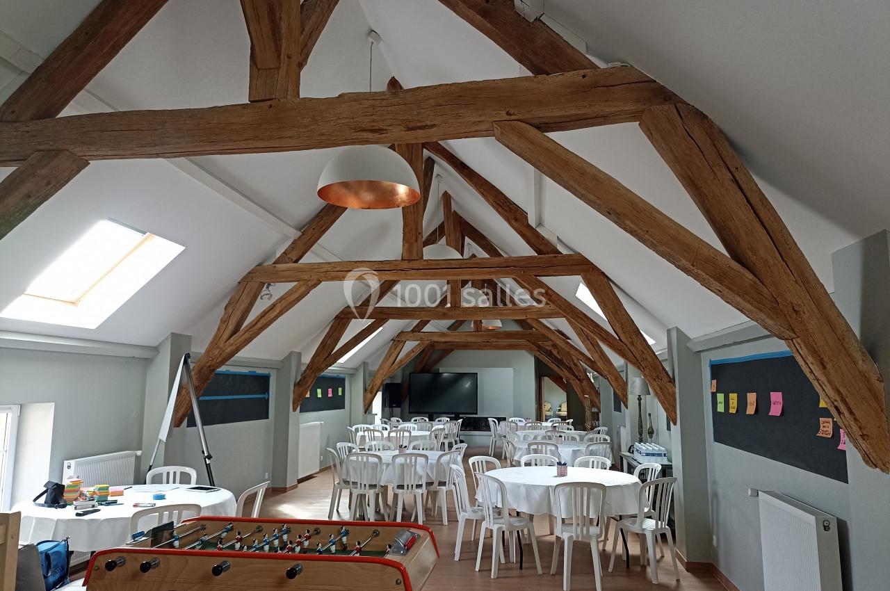 Salle lumineuse sous combles avec poutres apparentes, tables rondes, chaises blanches et baby-foot au premier plan.