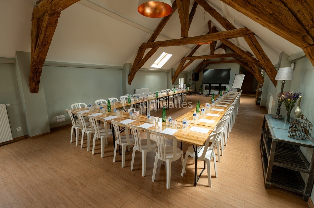 Salle de réunion lumineuse avec poutres apparentes, tables en U, chaises blanches et matériel de conférence.