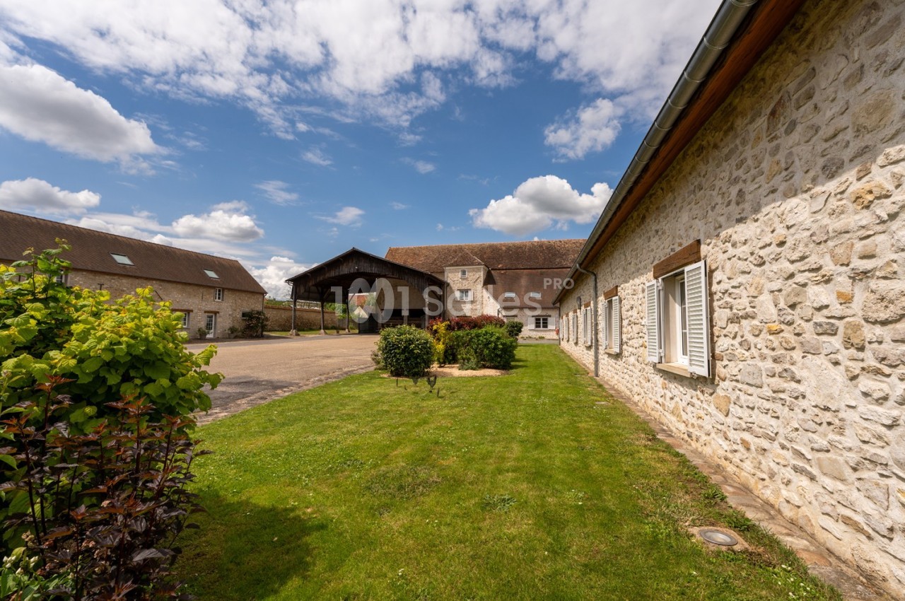 Cour d'une ferme avec bâtiments en pierre, pelouse bien entretenue et ciel partiellement nuageux.