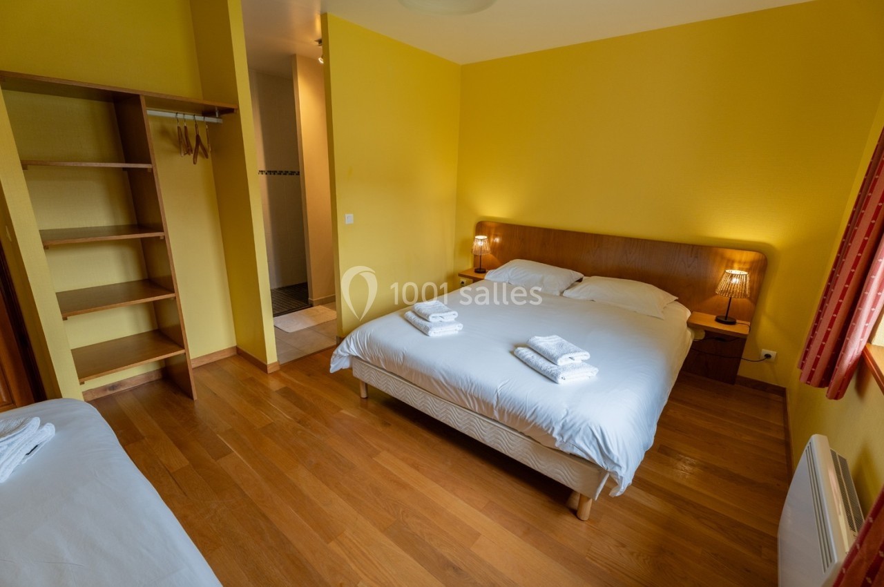 Chambre avec lit double, lit simple, parquet, murs jaunes, étagères ouvertes et salle de bain attenante visible.