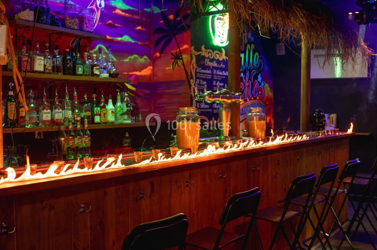 Bar en bois avec une rangée de flammes sur le comptoir, entouré de chaises pliantes et décor tropical lumineux.