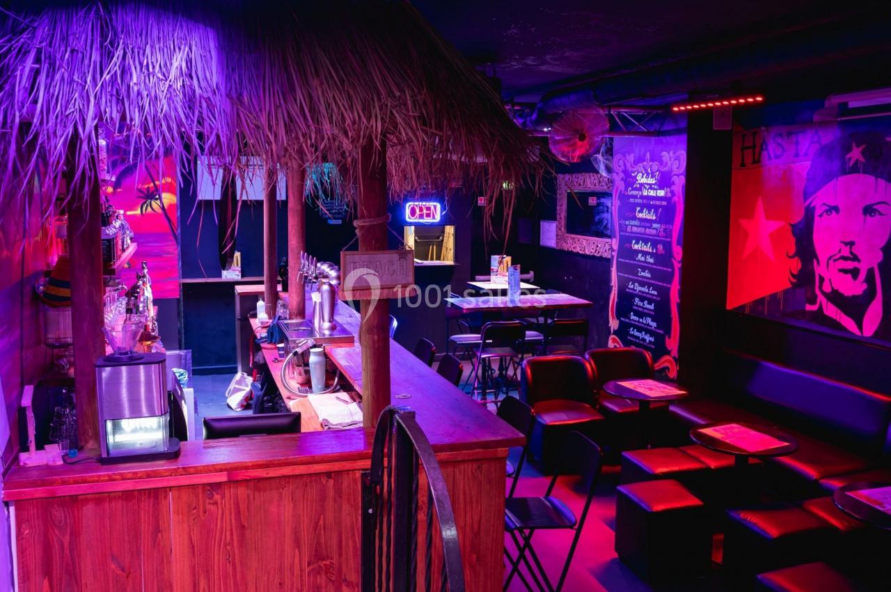 Bar avec comptoir en bois, chaises noires, éclairage coloré et décoration murale à thème tropical et révolutionnaire.