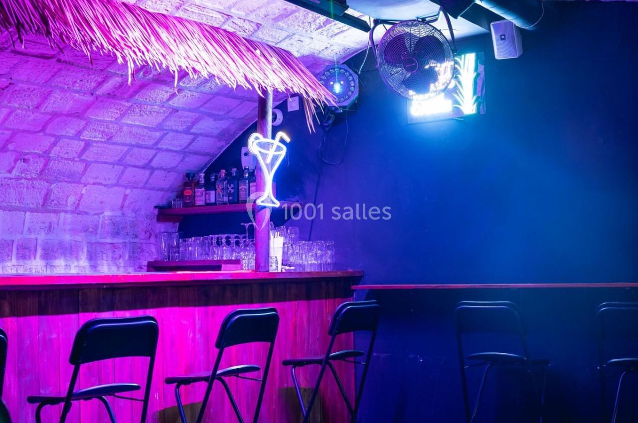 Bar avec comptoir en bois, tabourets alignés, éclairage violet et néon en forme de verre dans une ambiance tamisée.