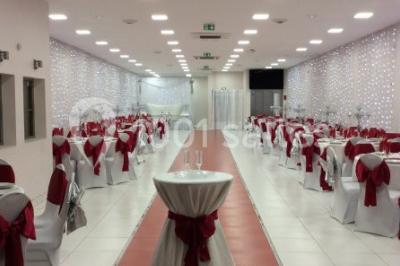 Salle décorée pour un événement avec tables rondes, nappes blanches, chaises ornées de nœuds argentés et dorés, éclairage…