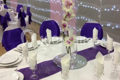 Salle décorée pour un événement avec tables rondes, nappes blanches, chaises ornées de nœuds argentés et dorés, éclairage…