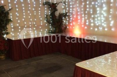 Salle décorée pour un événement avec tables rondes, nappes blanches, chaises ornées de nœuds argentés et dorés, éclairage…