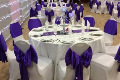 Salle décorée pour un événement avec tables rondes, nappes blanches, chaises ornées de nœuds argentés et dorés, éclairage…