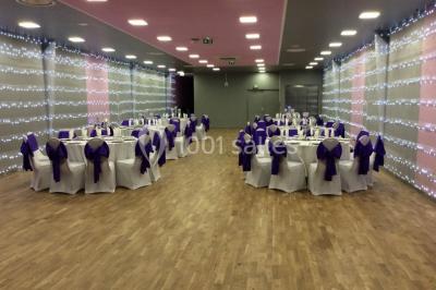 Salle décorée pour un événement avec tables rondes, nappes blanches, chaises ornées de nœuds argentés et dorés, éclairage…