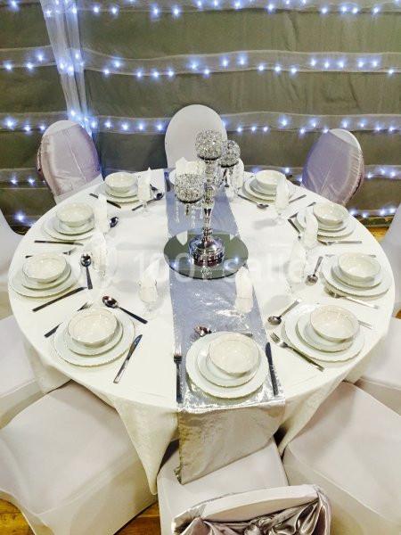 Table ronde décorée avec une nappe blanche, couverts disposés pour huit personnes et centre de table argenté.