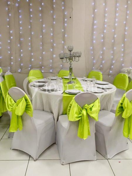 Table ronde dressée avec nappes blanches, chaises décorées de nœuds verts, chandelier central et guirlandes lumineuses en…