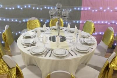 Salle décorée pour un événement avec tables rondes, nappes blanches, chaises ornées de nœuds argentés et dorés, éclairage…