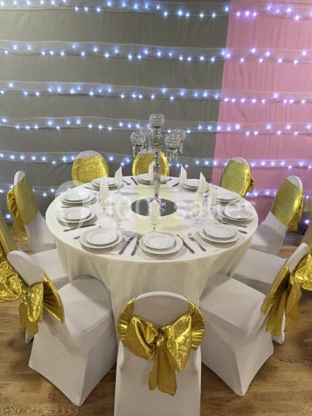 Table ronde décorée pour un événement, avec nappes blanches, serviettes pliées, chaises ornées de nœuds dorés et centre de…