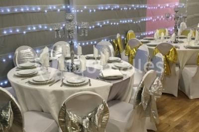 Salle décorée pour un événement avec tables rondes, nappes blanches, chaises ornées de nœuds argentés et dorés, éclairage…