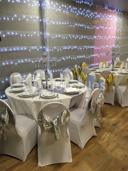 Salle décorée pour un événement avec tables rondes, nappes blanches, chaises ornées de nœuds argentés et dorés, éclairage…