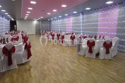 Salle décorée pour un événement avec tables rondes, nappes blanches, chaises ornées de nœuds argentés et dorés, éclairage…