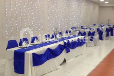 Salle décorée pour un événement avec tables rondes, nappes blanches, chaises ornées de nœuds argentés et dorés, éclairage…