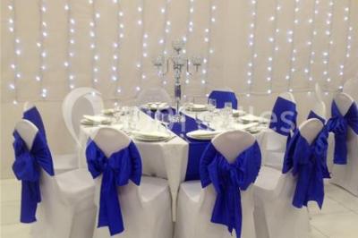 Salle décorée pour un événement avec tables rondes, nappes blanches, chaises ornées de nœuds argentés et dorés, éclairage…