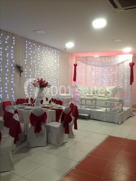 Salle décorée pour un événement avec des chaises blanches ornées de nœuds rouges, table dressée et canapé sur estrade.