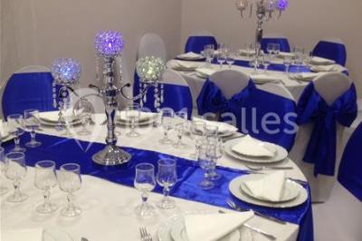 Salle décorée pour un événement avec tables rondes, nappes blanches, chaises ornées de nœuds argentés et dorés, éclairage…