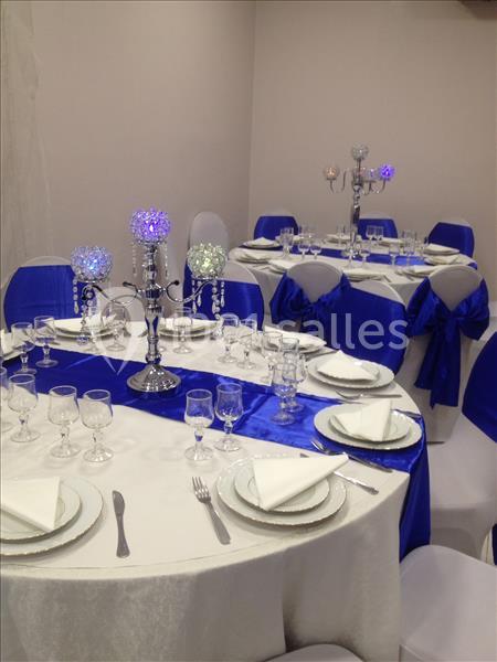 Tables rondes élégamment dressées avec nappes blanches, chemins de table bleus, serviettes pliées et chandeliers décoratifs.