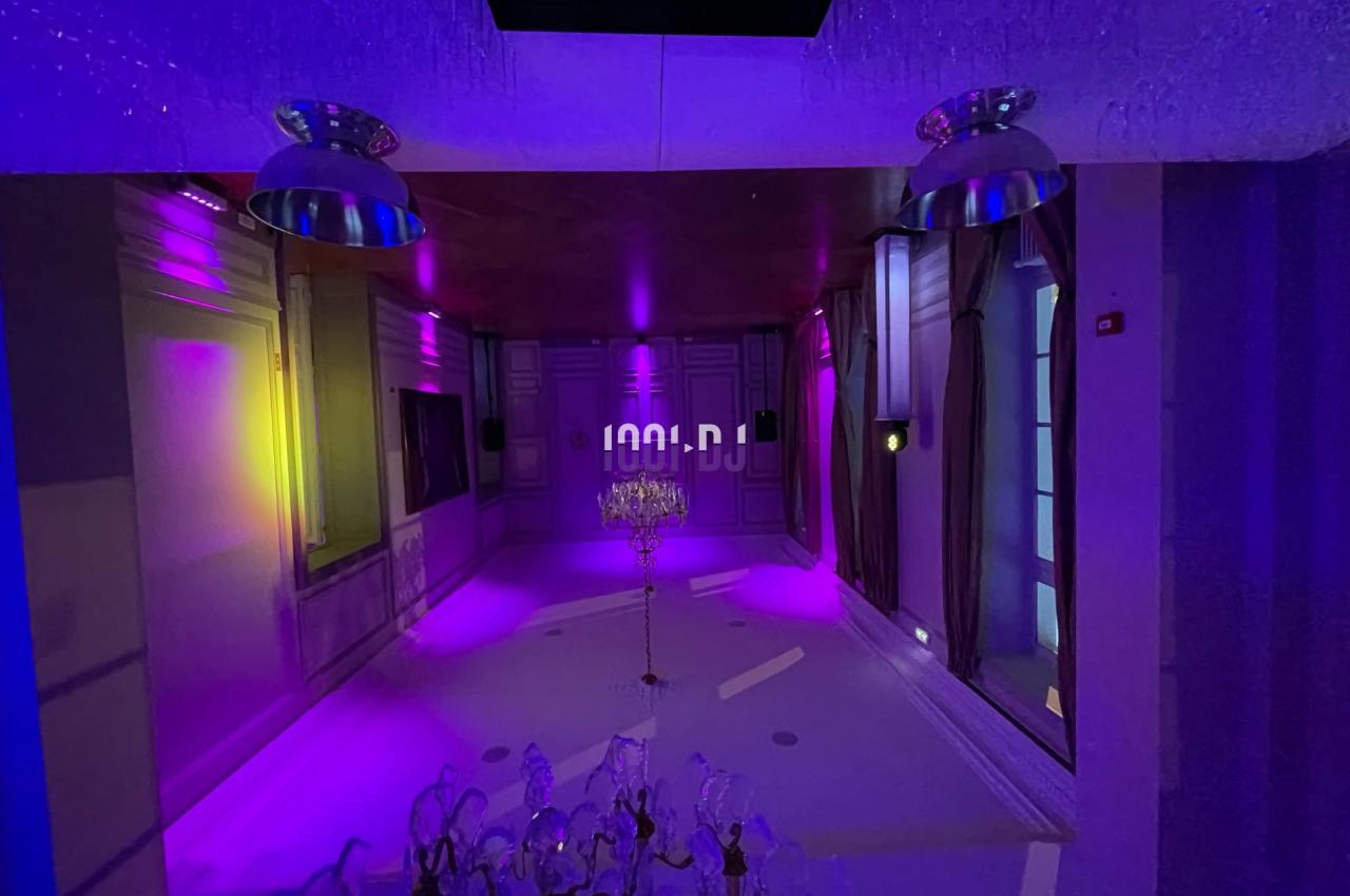 Salle éclairée par des lumières colorées violettes et jaunes, avec un lustre central et des murs ornés de moulures.