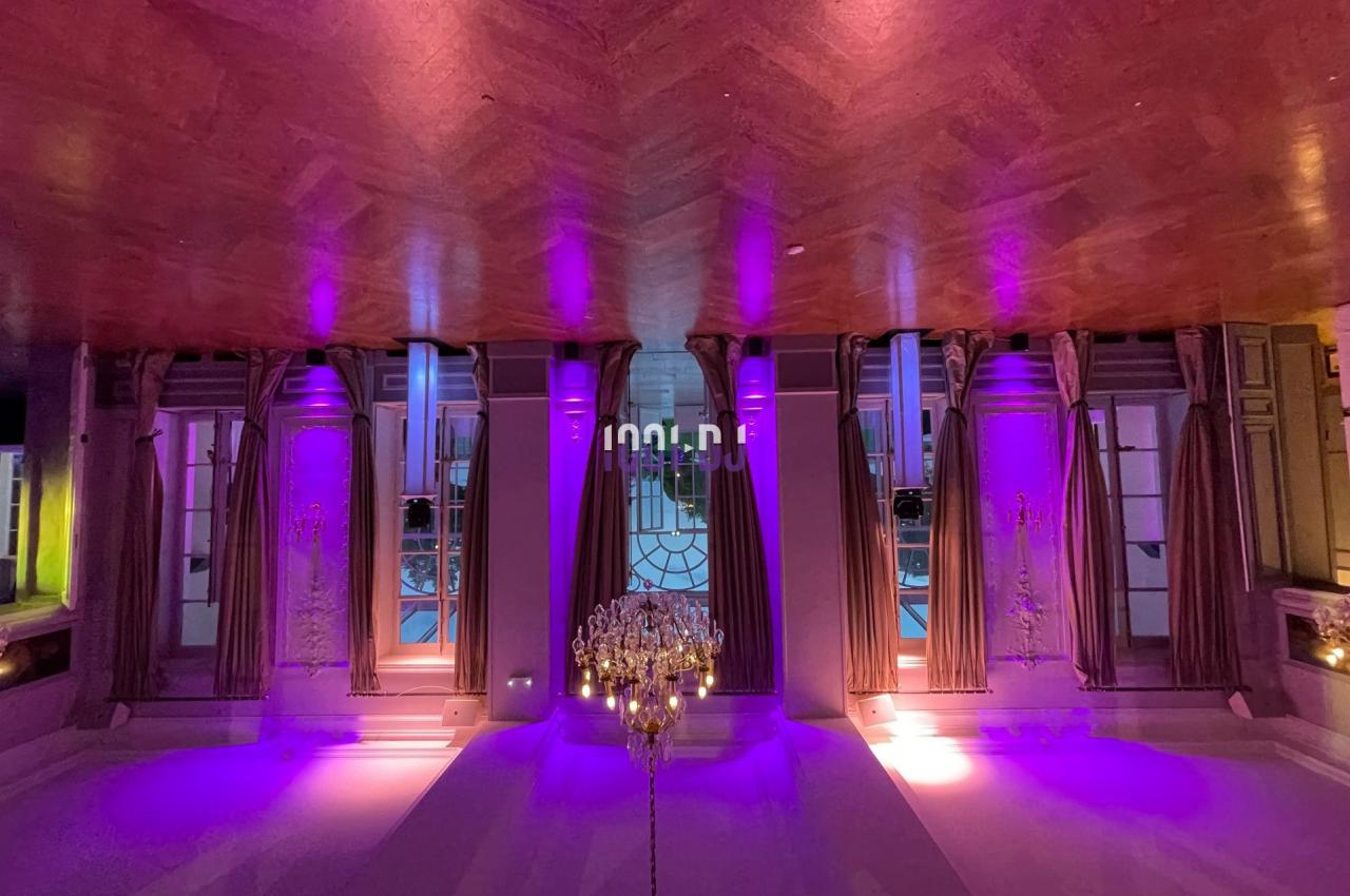 Salle élégante avec parquet, lustre central et éclairage violet mettant en valeur des fenêtres avec rideaux.