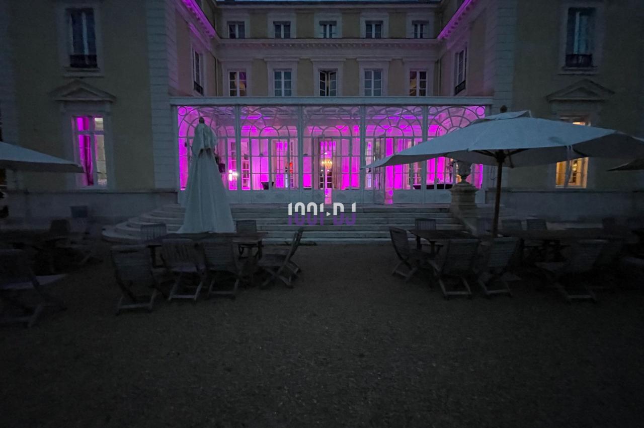 Façade d'un bâtiment éclairé en rose, avec terrasse aménagée de tables et parasols au premier plan, en soirée.