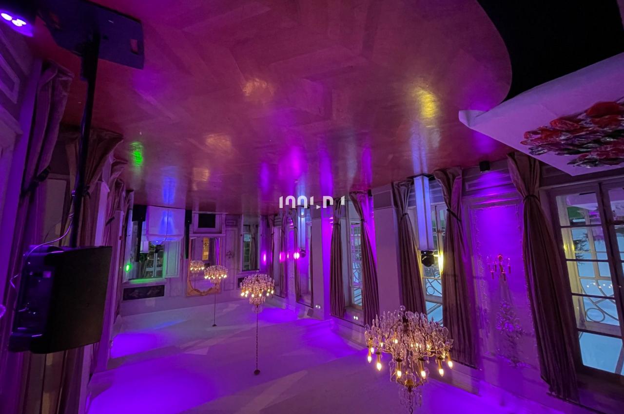 Salle élégante avec parquet, lustres et éclairage violet, entourée de grandes fenêtres et rideaux.