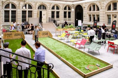 Terrain de pétanque en plein air avec chaises pliantes, parasol et espace ombragé entouré de verdure.