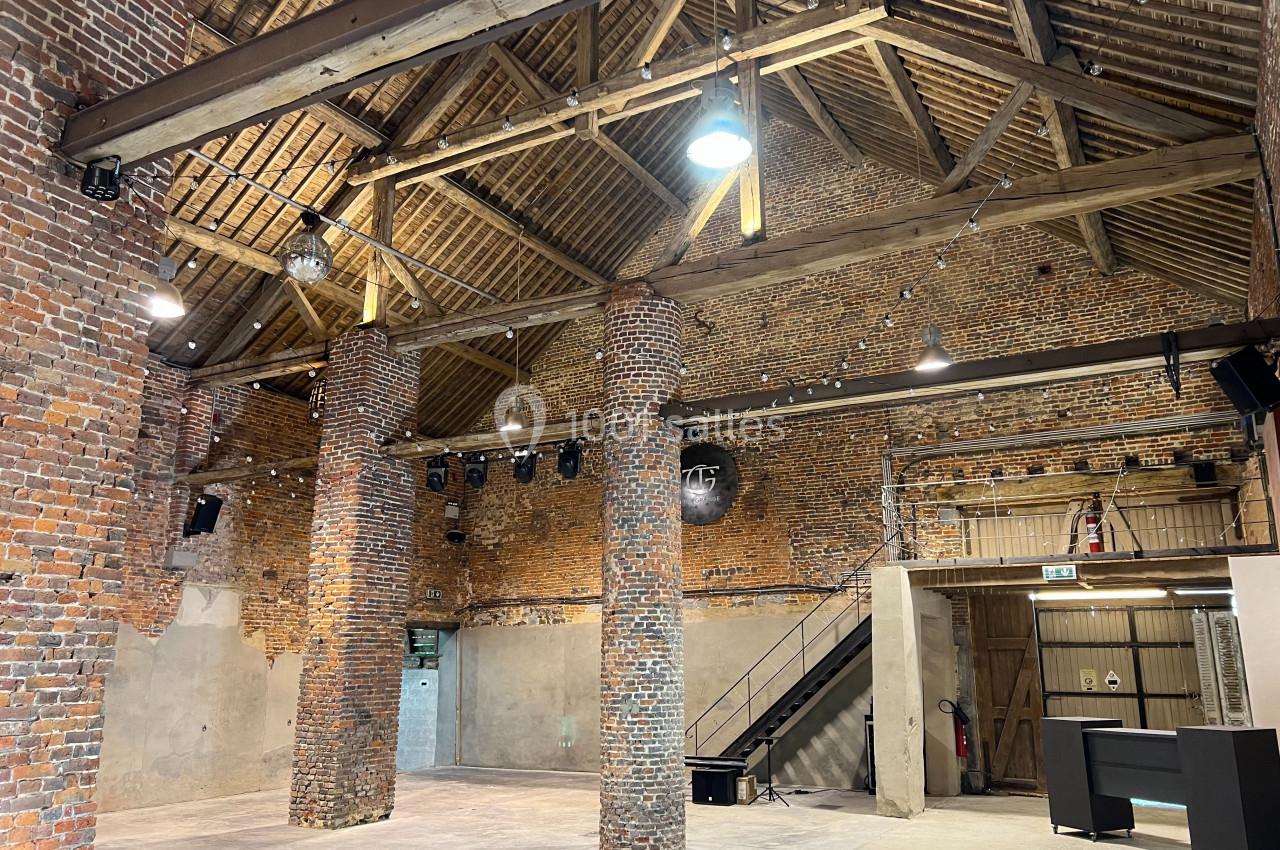 Intérieur d'une grande salle en briques avec poutres apparentes, colonnes, éclairage suspendu et escalier métallique.