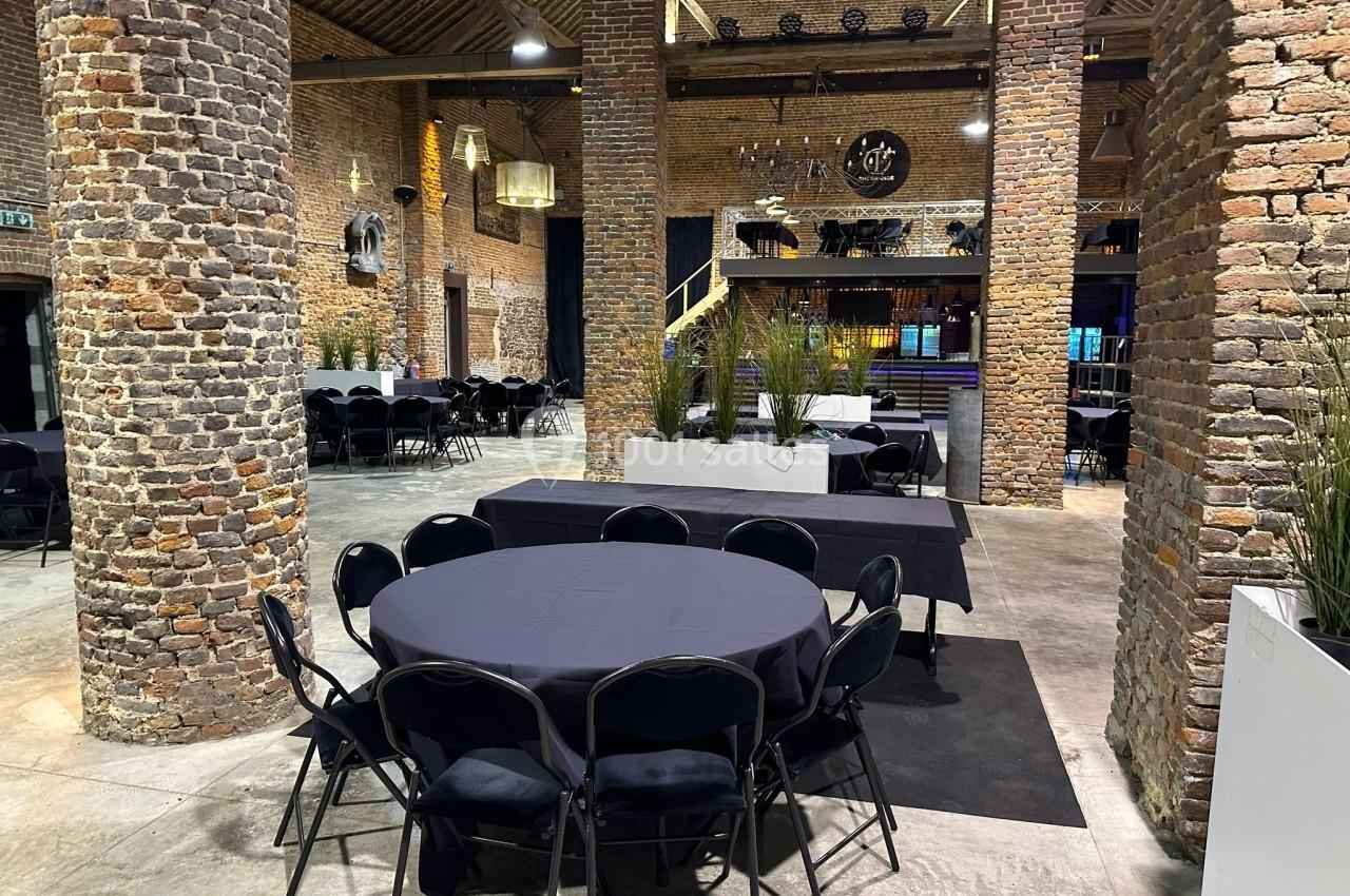 Salle en briques avec tables rondes et chaises noires, éclairage tamisé et mezzanine en arrière-plan.