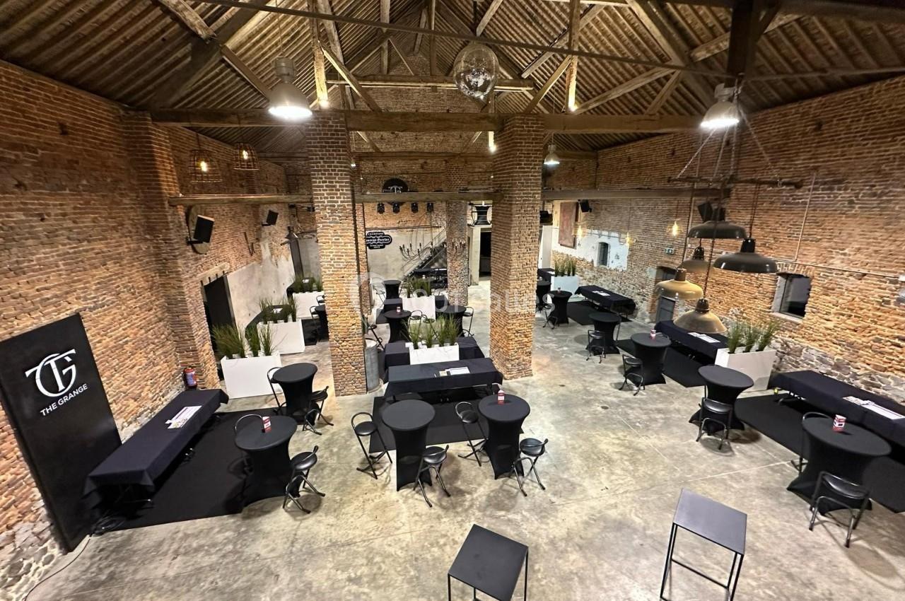 Salle en briques avec charpente apparente, aménagée avec des tables hautes et des chaises pour un événement.