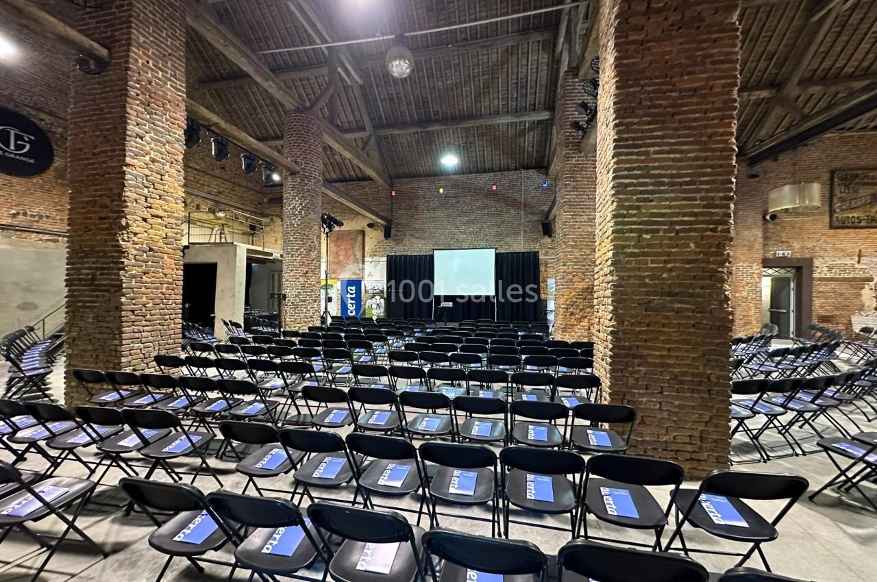 Salle en briques avec des rangées de chaises noires alignées face à une scène équipée d'un écran de projection.