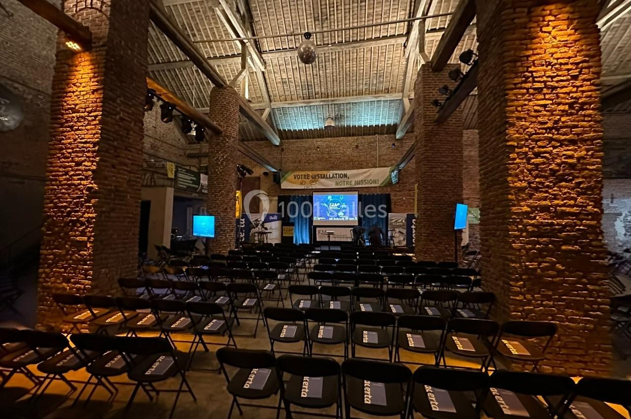 Salle de conférence en briques avec des rangées de chaises noires vides, un écran et une scène éclairée à l'arrière-plan.