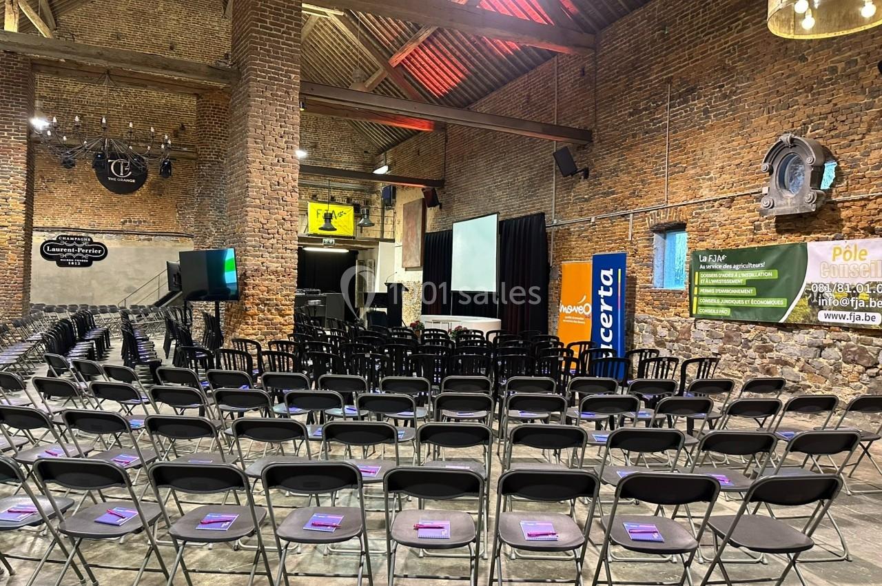 Salle en briques avec des rangées de chaises noires alignées, écran de projection et affiches promotionnelles au fond.