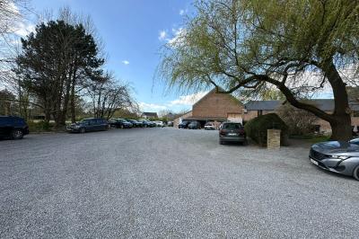 Parking en gravier entouré d'arbres et de bâtiments en briques sous un ciel partiellement nuageux.