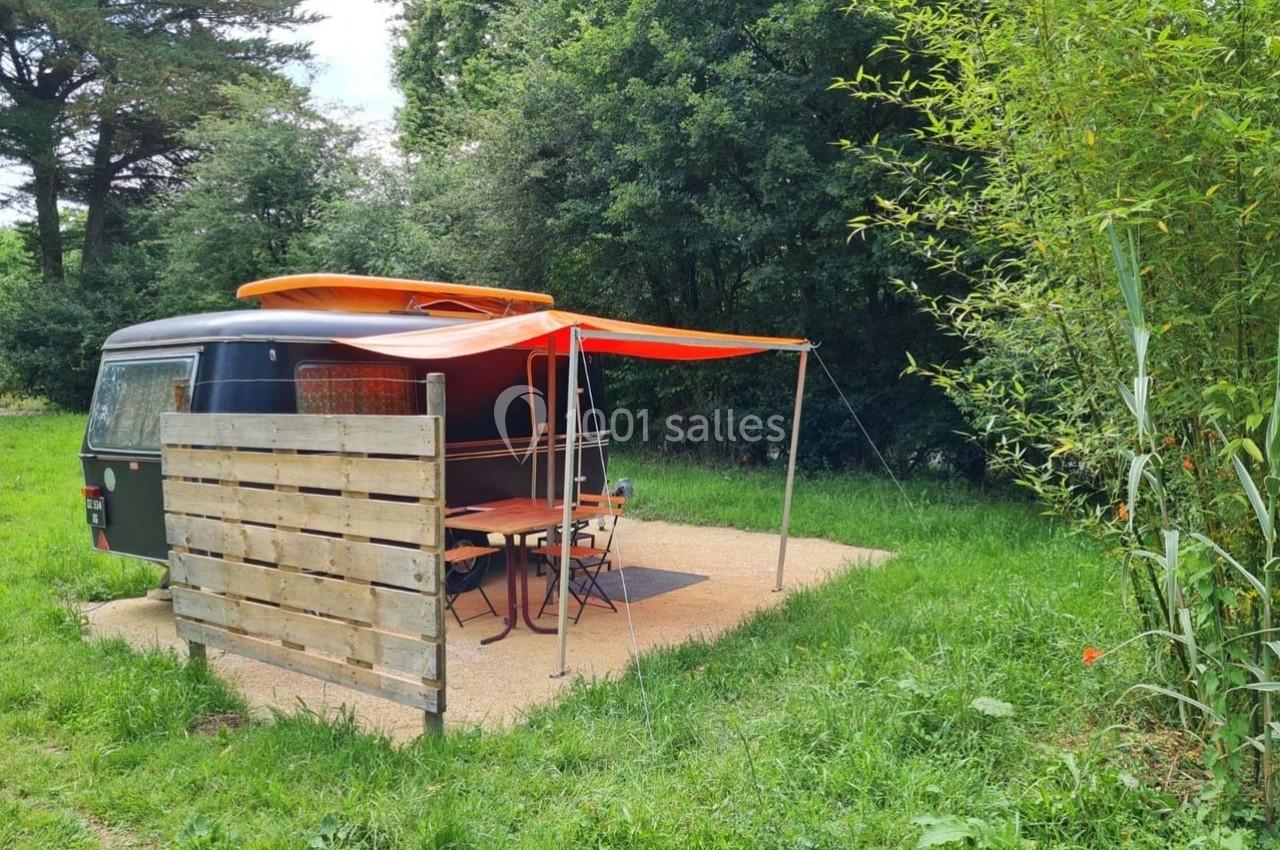 Caravane noire avec auvent orange installée sur un emplacement herbeux entouré d'arbres et de bambous.