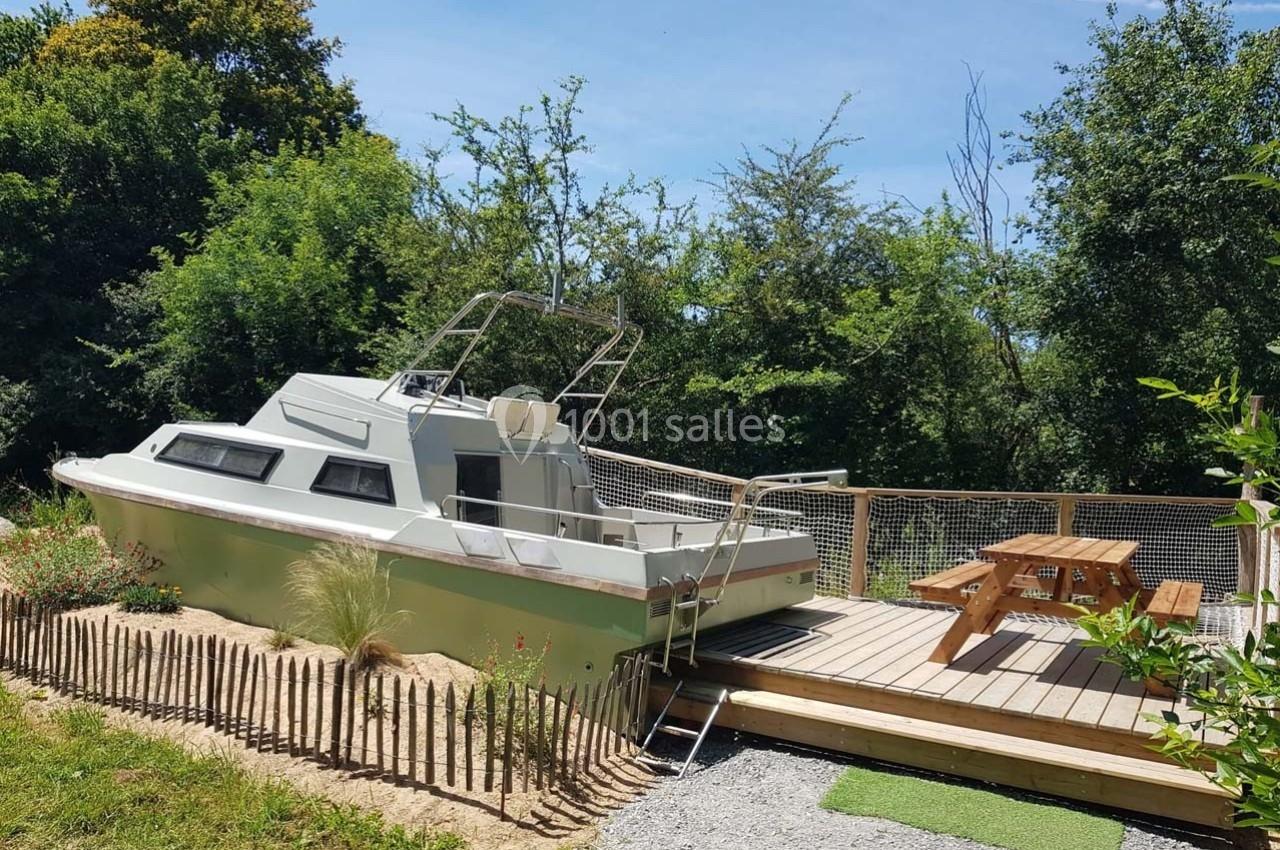 Bateau aménagé en hébergement insolite sur une terrasse en bois, entouré de verdure et d'une clôture en bois.